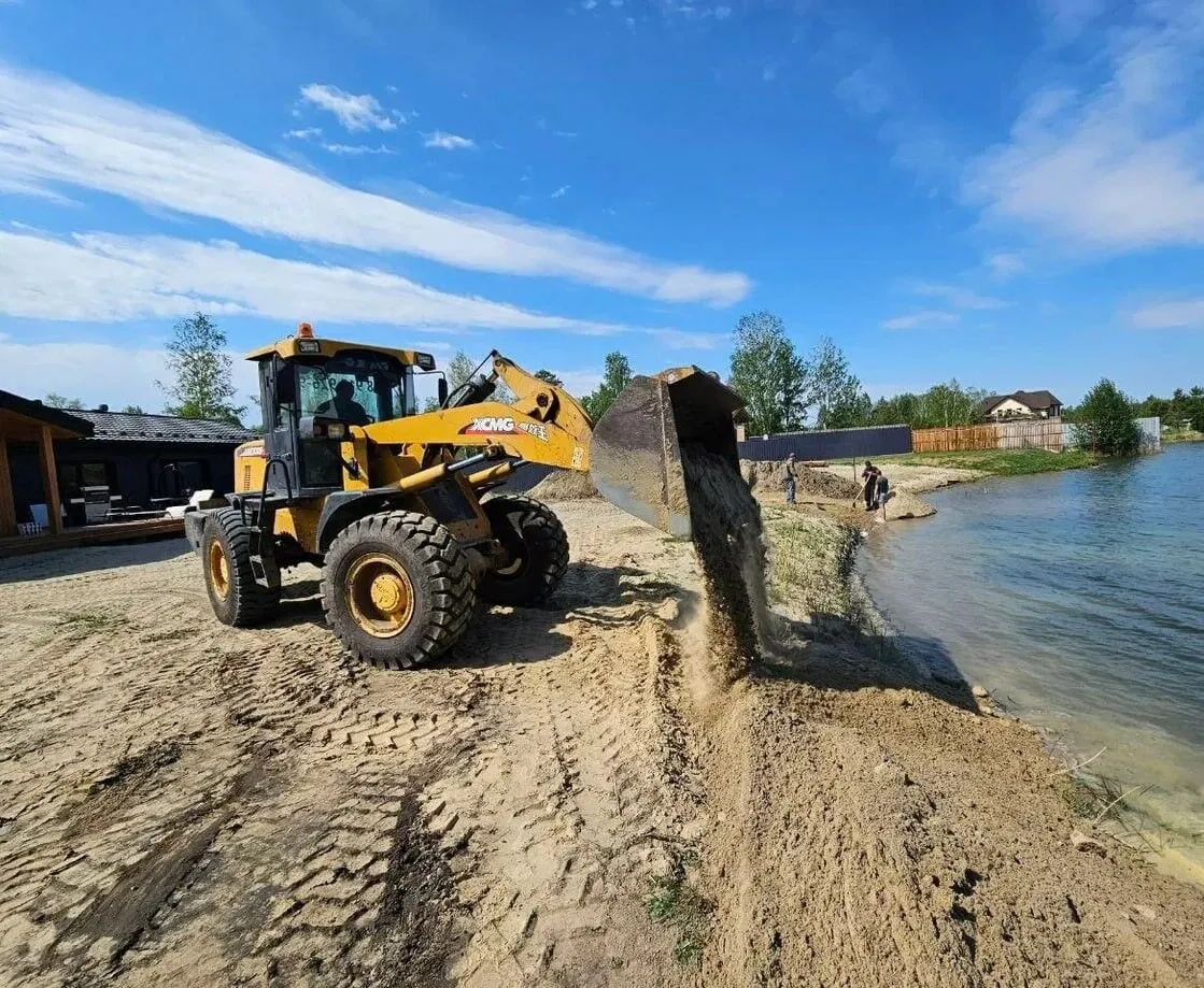 Фронтальный погрузчик высыпает песок на берегу водоёма.