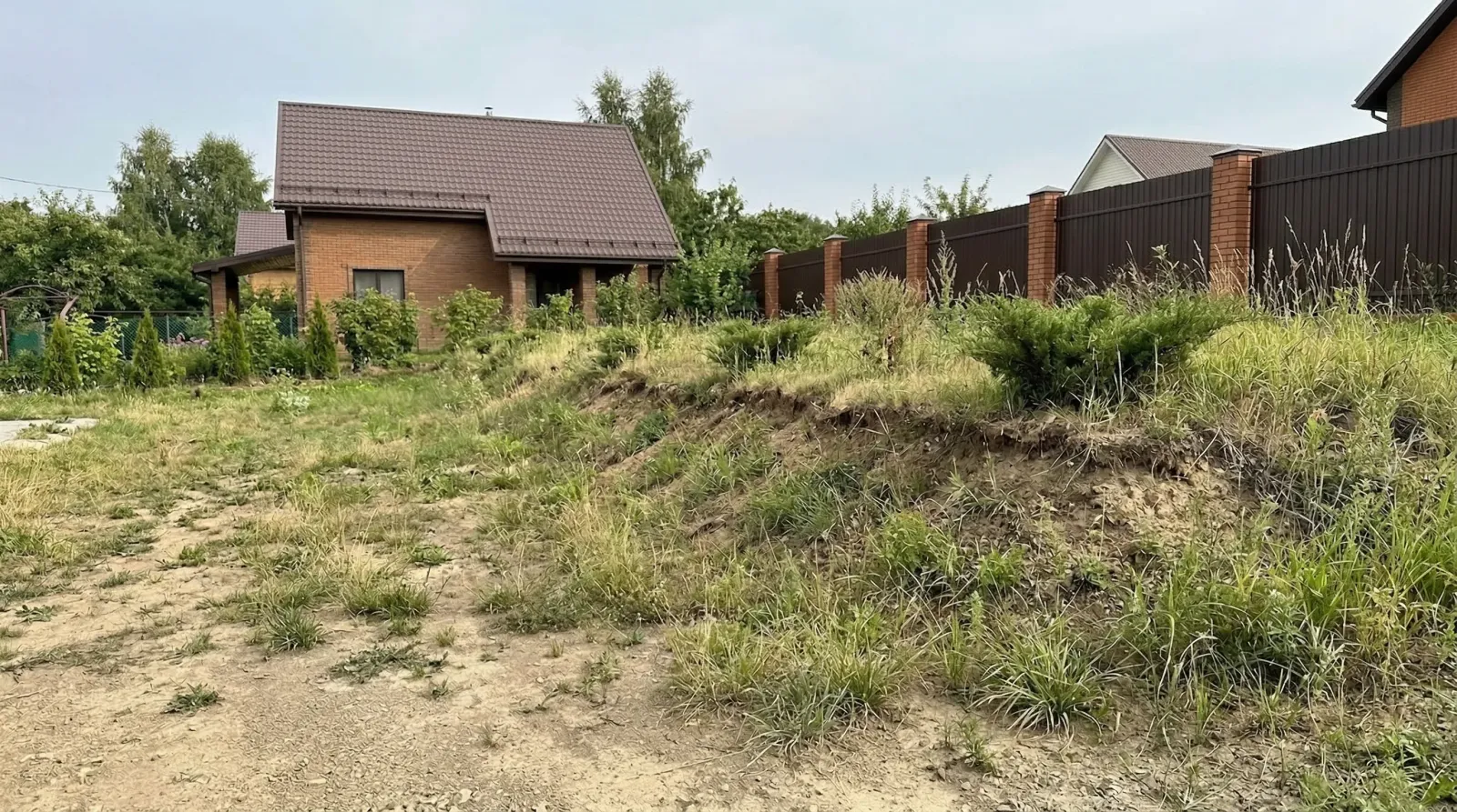 Загородный кирпичный дом за заросшим травой неровным участком.