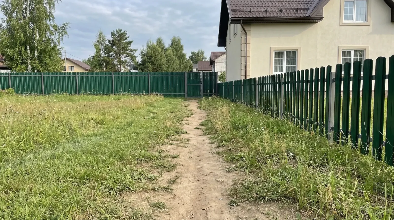 Заросший травой участок с тропинкой вдоль зелёного забора.