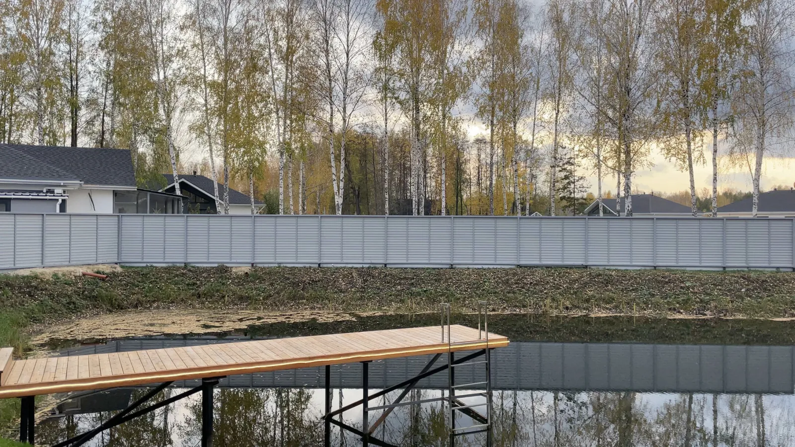Деревянный причал у пруда на фоне берёз и домов.