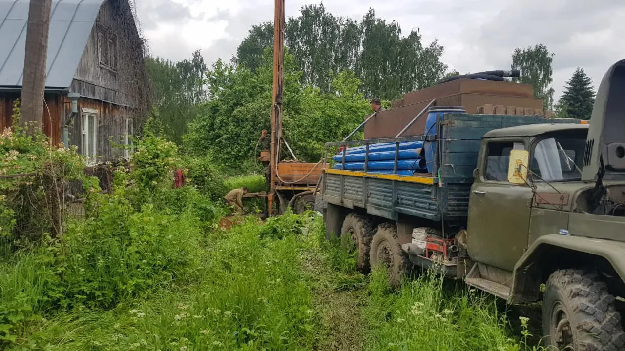 Буровая установка работает возле деревенского дома среди зелени.